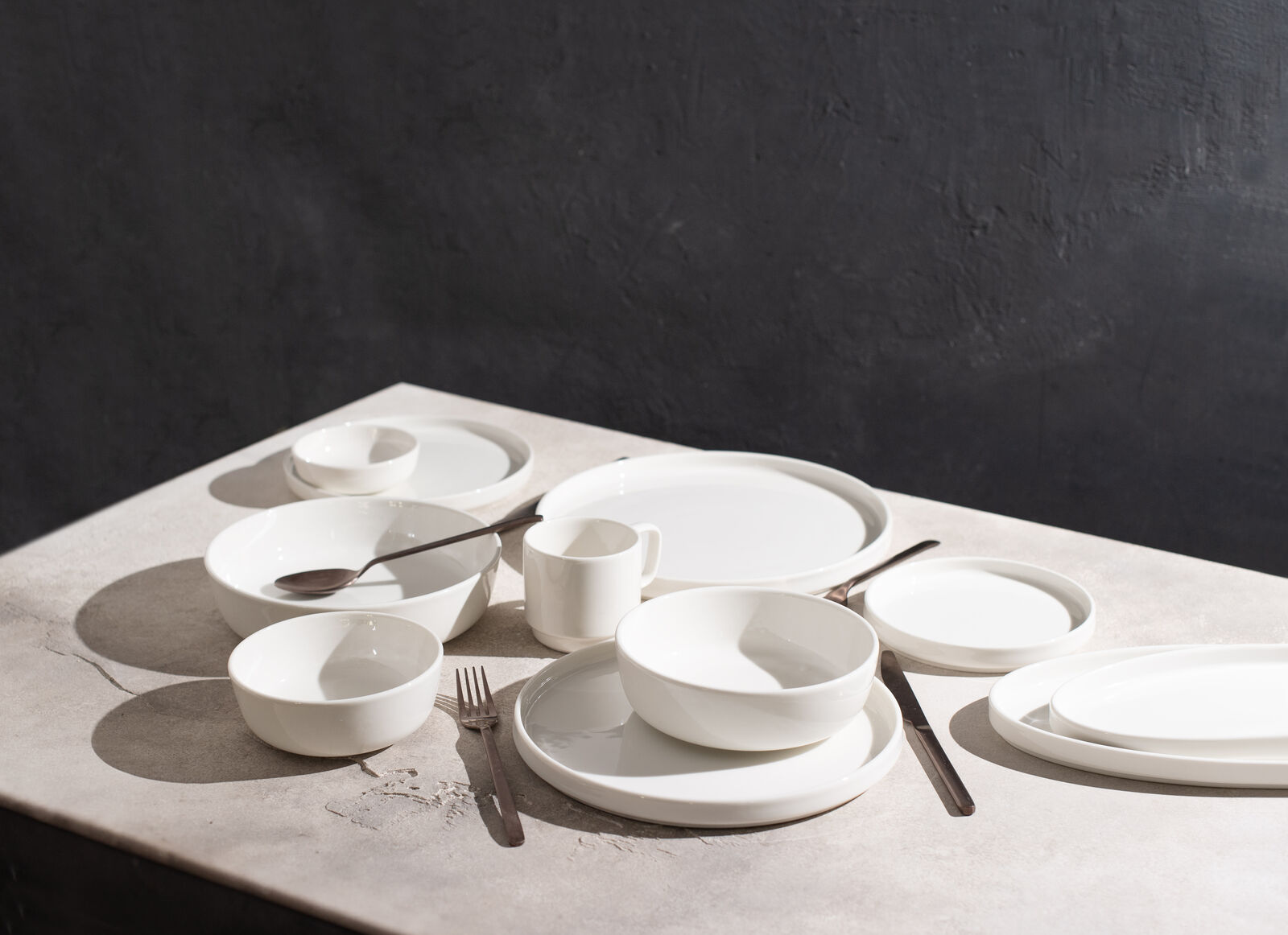 White tableware set on a light stone table, including plates, bowls, and utensils against a dark gray background, creating a minimalist feel.
