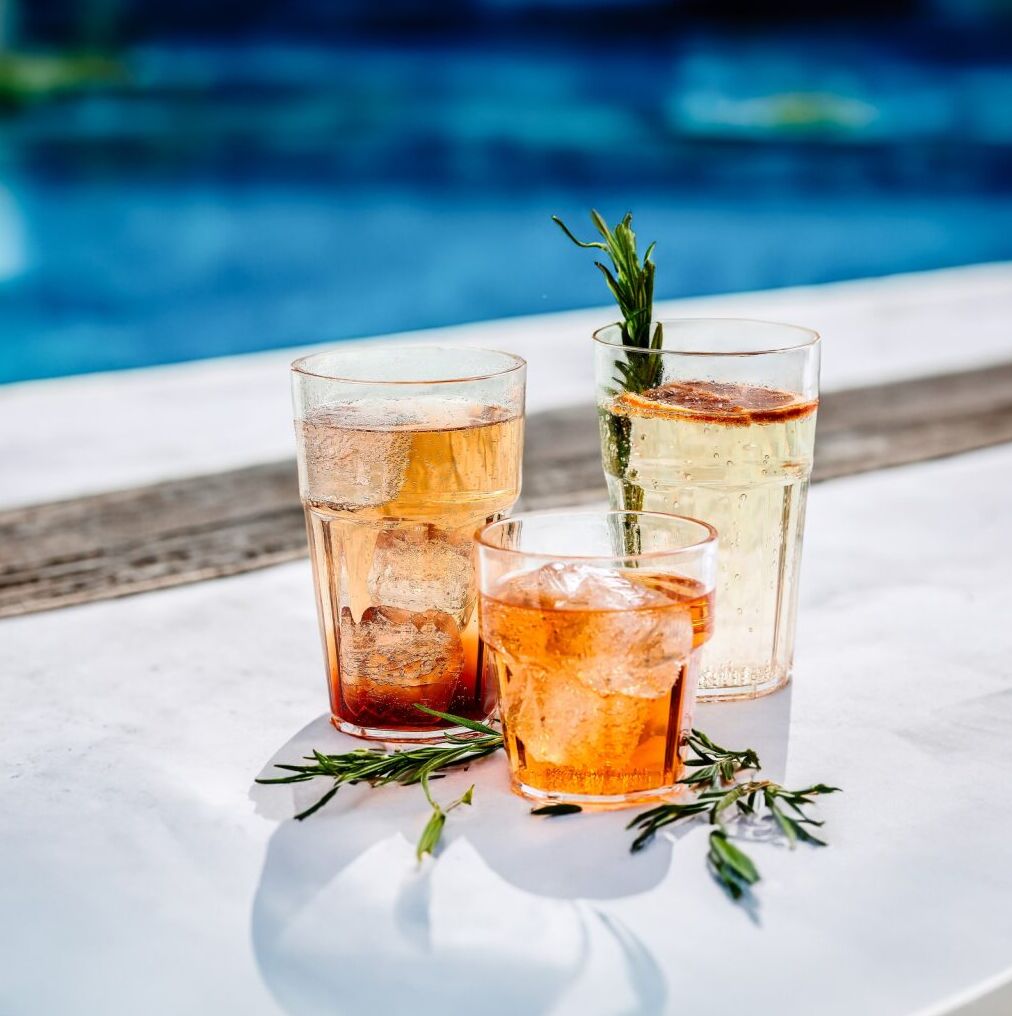 Three colorful drinks with ice and herbs on a white table by a pool, sunlight creating a refreshing summer vibe.