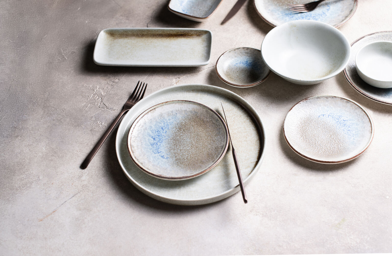 Ceramic plates and bowls with blue and beige speckled patterns on a textured surface. Fork and knife placed diagonally on the largest plate.