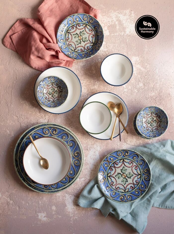 Ceramic bowls and plates with intricate floral designs on a textured surface with pink and teal fabrics. Two gold spoons. Mood: elegant.