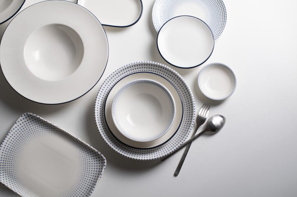 White plates and bowls with blue dotted patterns and silver cutlery on a white surface. Neatly arranged, creating a clean, elegant look.