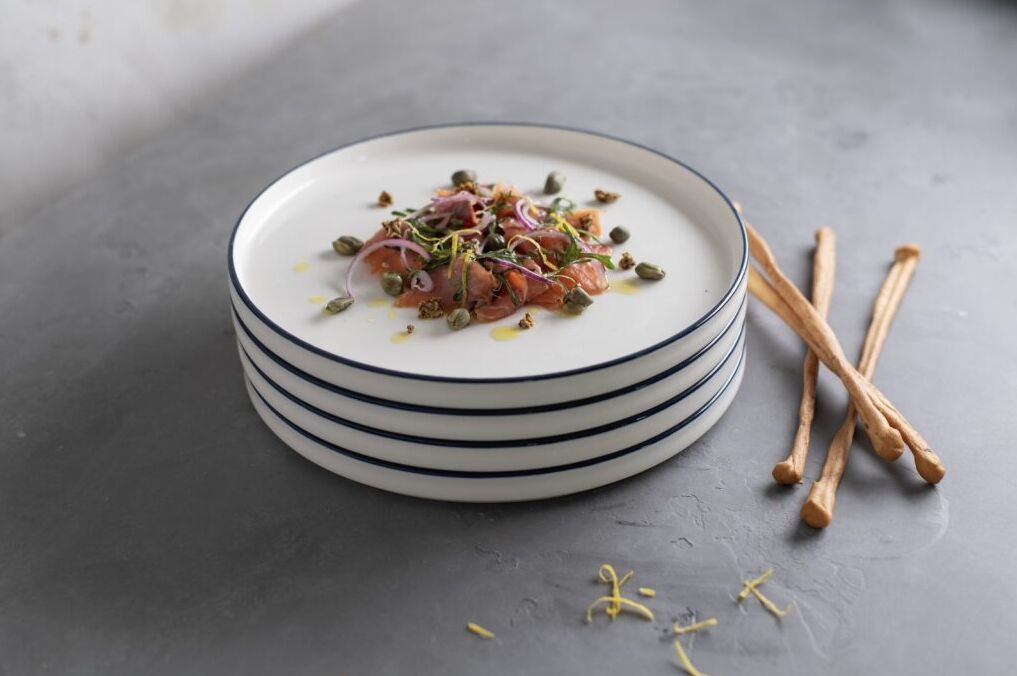 Stacked white plates with a dish of salmon, capers, and onions, garnished with herbs. On a gray table with breadsticks beside them.