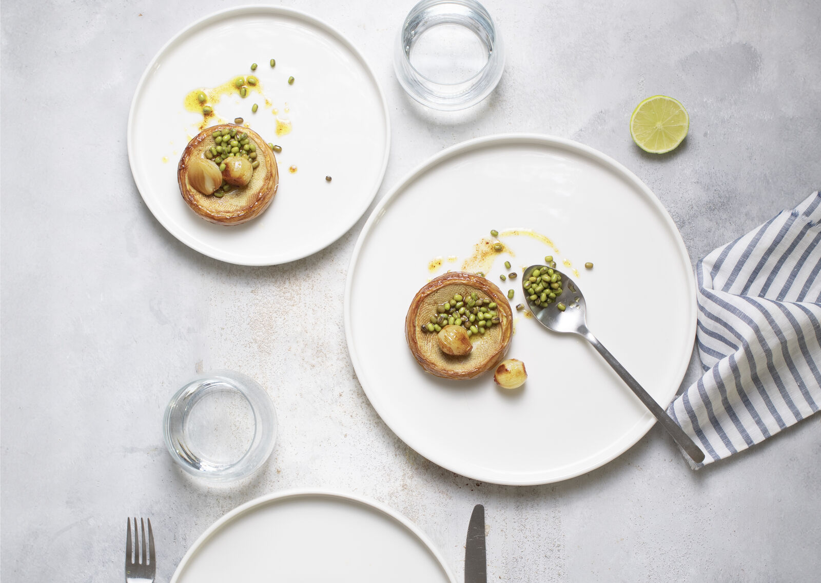 Two white plates with savory pastries topped with green peas and onions. A fork, spoon, glass, half lime, and striped napkin on gray surface.