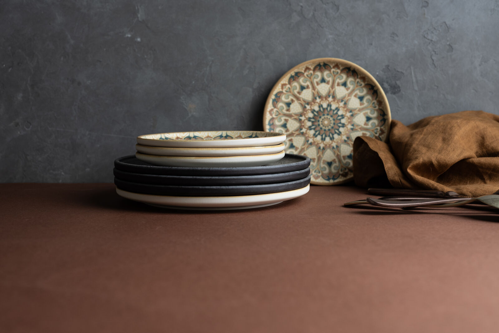 Stacked plates with black and patterned designs on a brown surface, gray textured background, brown cloth, and cutlery beside it.
