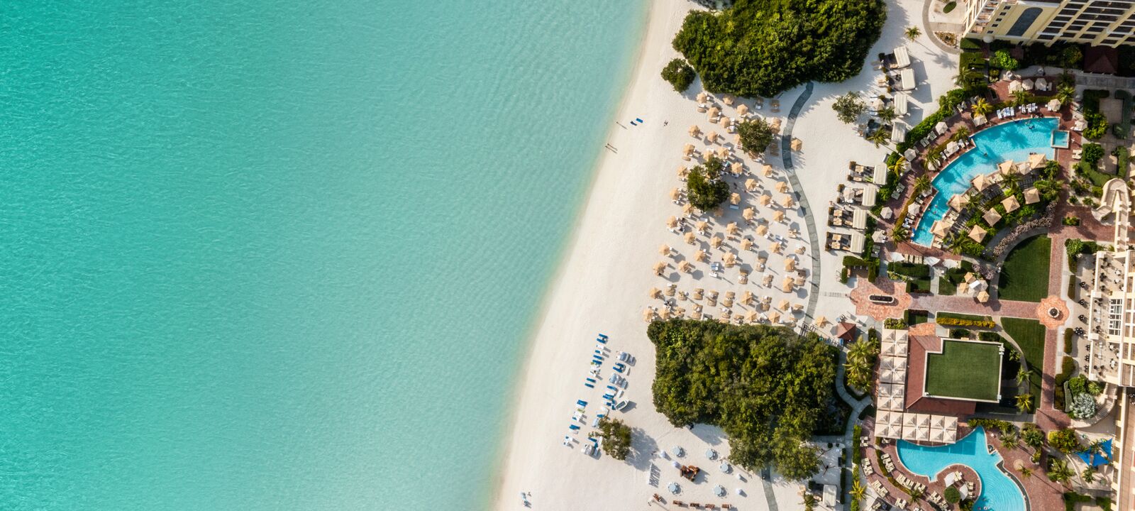 Aerial view of a beach resort with turquoise water, white sand, sun loungers, and umbrellas. Lush greenery and pool area visible. Bright, sunny day.