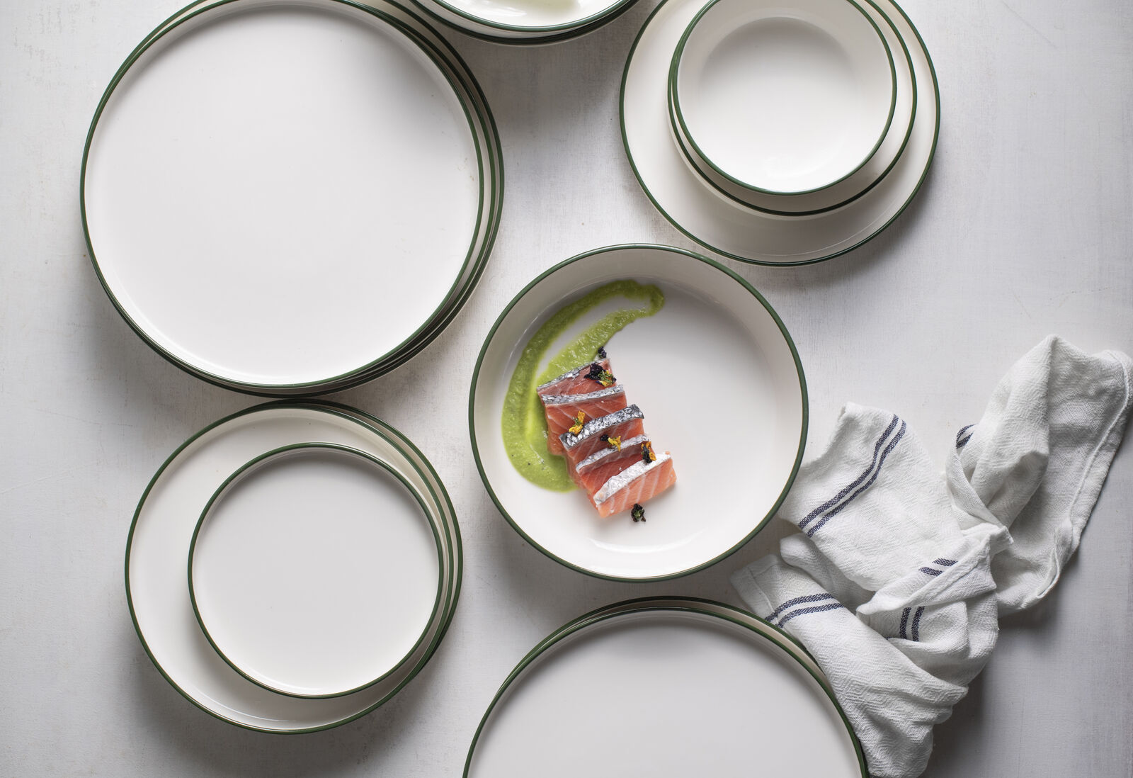 White plates with green rims on a table, one with sliced salmon and green sauce. A white cloth with stripes lies beside them. Elegant setting.