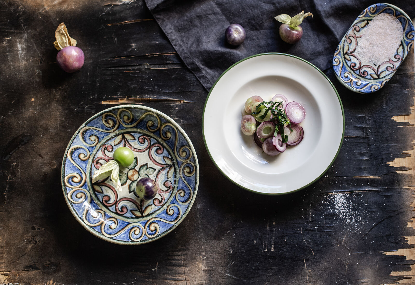Colorful plates with sliced onions and tomatoes on a rustic wooden table. Decorative patterns and dark cloth add an artistic touch.