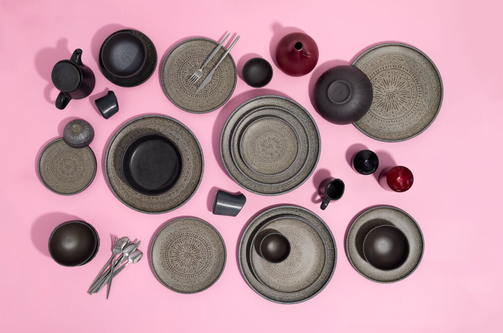 Black patterned dinnerware and cutlery arranged on a pink background. Includes plates, cups, and vases creating a balanced design.